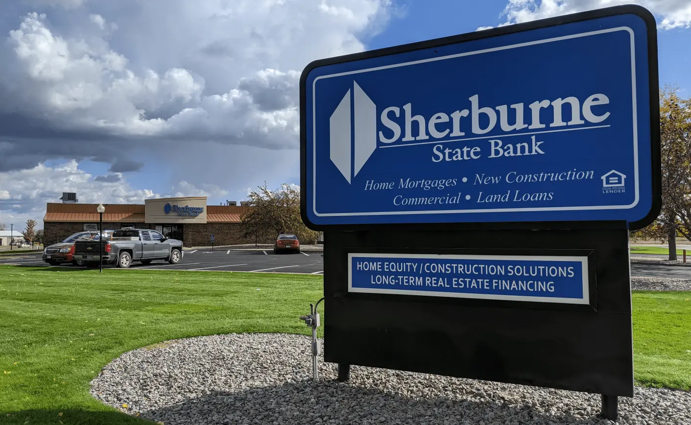 A blue sign for Sherburne State Bank stands on gravel, advertising home loans. It's set against a cloudy sky and a nearby building with parked cars.